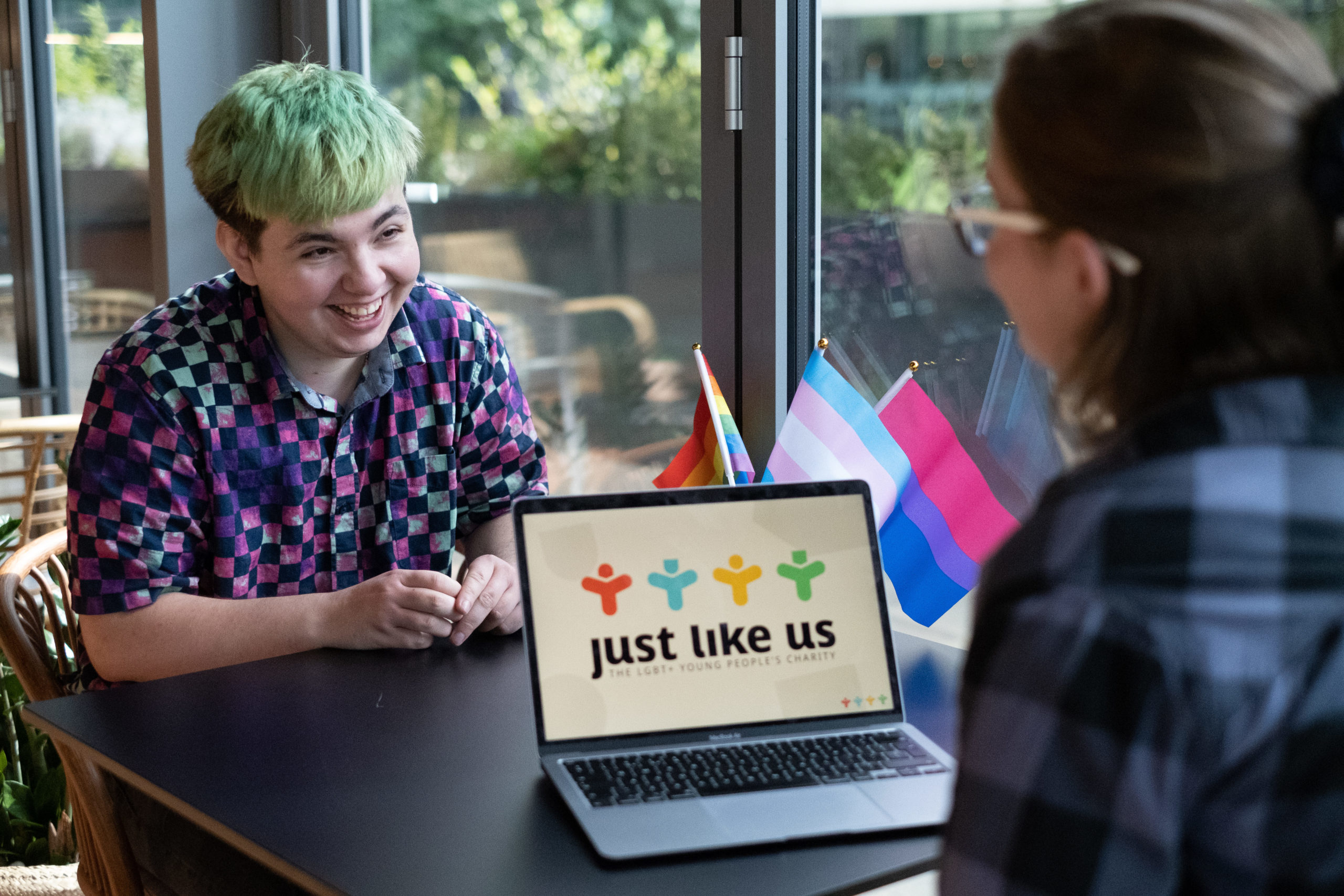 Two LGBT+ young people chatting and laughing, with Just Like Us on their laptop screen