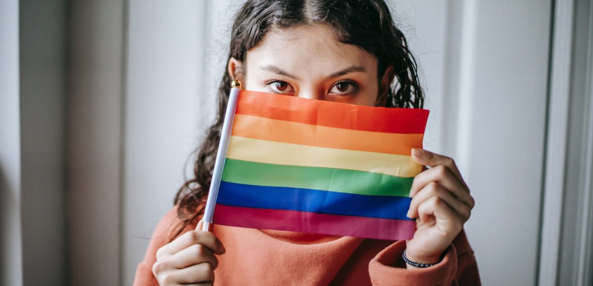 A girl, looking into the camera, stands with a Pride flag in front of her face