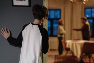 A young person stands in a house hallway, looking into the living room where parents are arguing