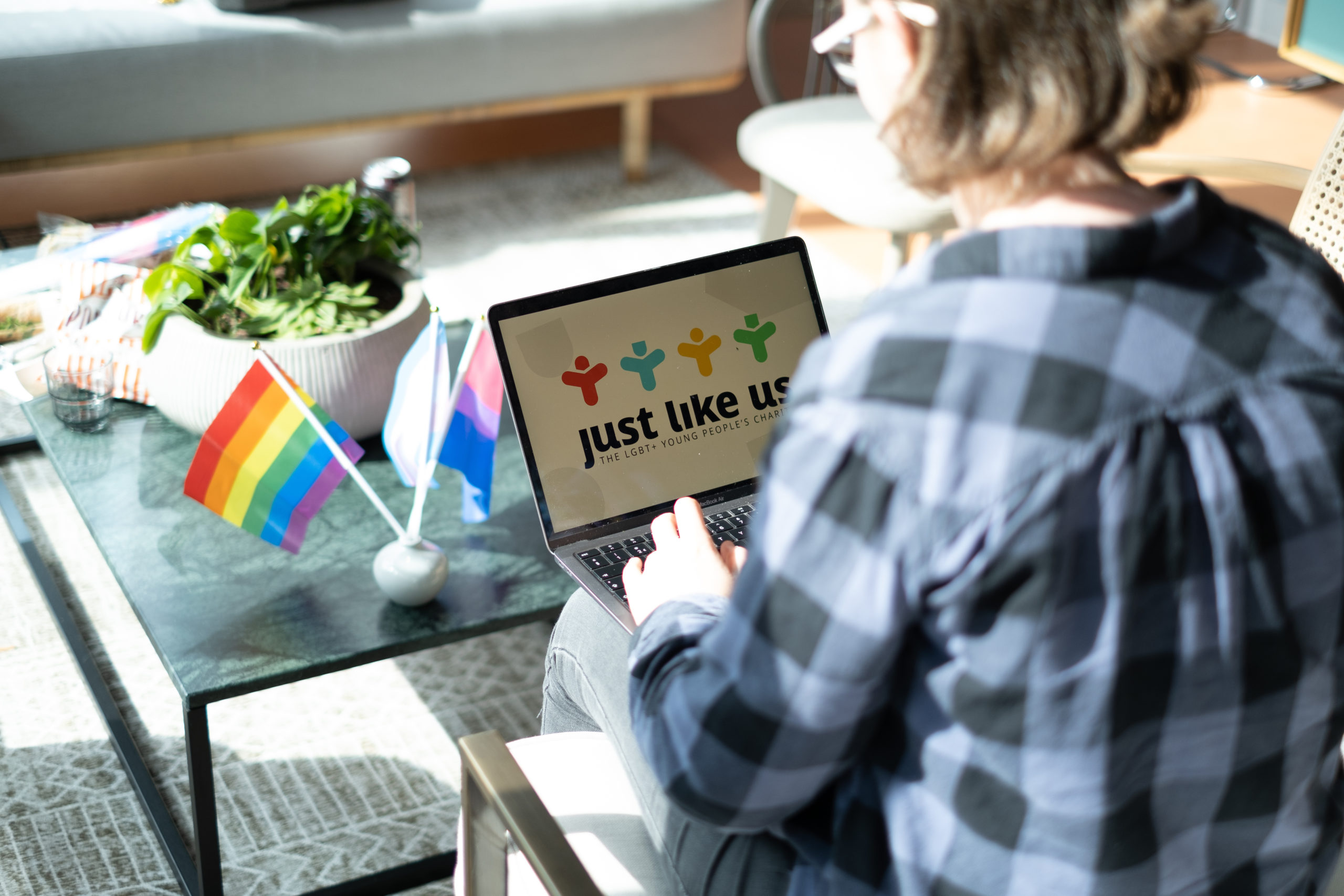 Get in touch: A person on a laptop, which has Just Like Us' logo on the screen, sat next to Pride flags