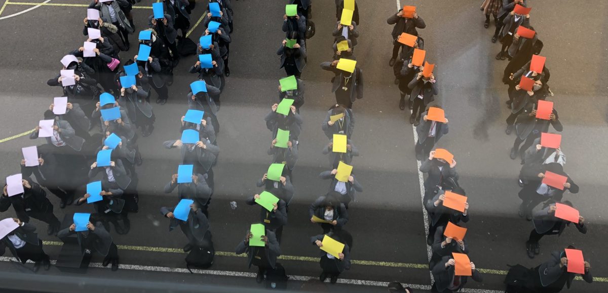 A photo from School Diversity Week celebrations taken from above of around 75 pupils in a playground, each holding up a different coloured piece of paper to represent the Pride flag