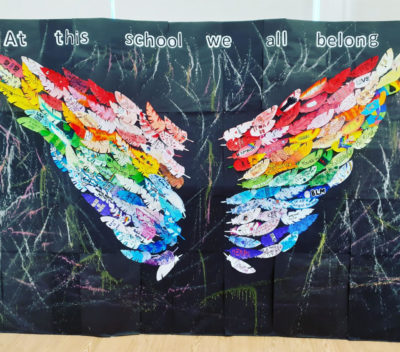 Photo of a school's School Diversity Week display, which says 'At this school we all belong' and has wings made of our rainbow-coloured feathers with messages in inclusion written on them