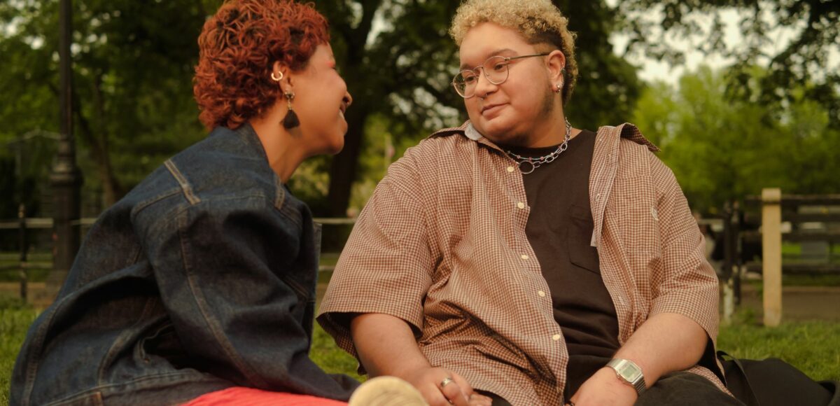 A trans couple smile at each other while sitting in the park