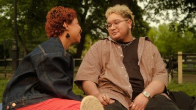 A trans couple smile at each other while sitting in the park