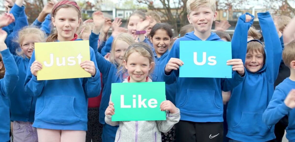 Primary school children hold signs that read Just Like Us in a playground - a screenshot from the School Diversity Week video
