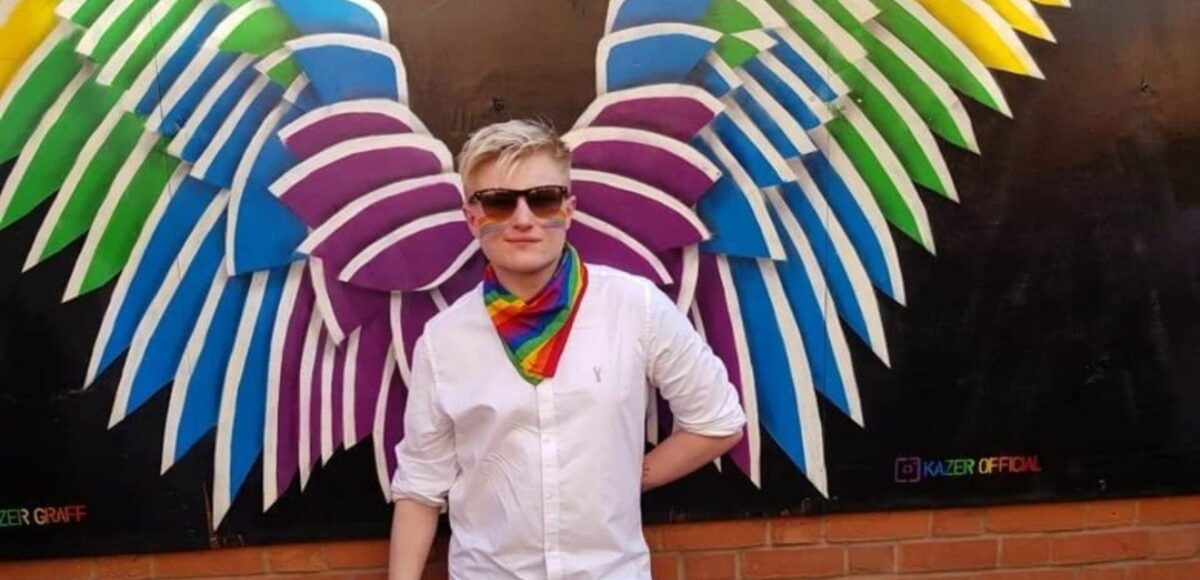 Just Like Us ambassador Sam Harvey stands in front of a pair of rainbow wings painted on a wall